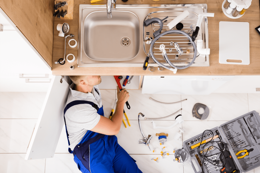 A plumber on the floor uses an orange wrench to fix the unseen pipe of a sink with their toolbox on the floor next to them