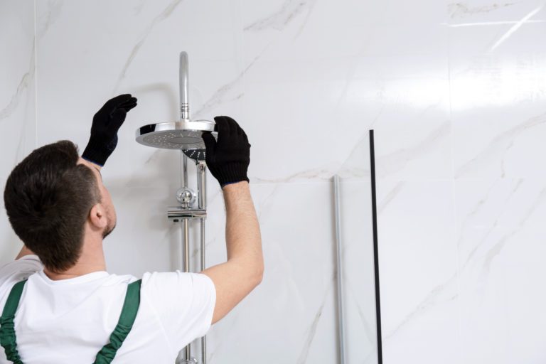 A plumber wearing gloves as he inspects a shower head in a bathroom with while marble walls.