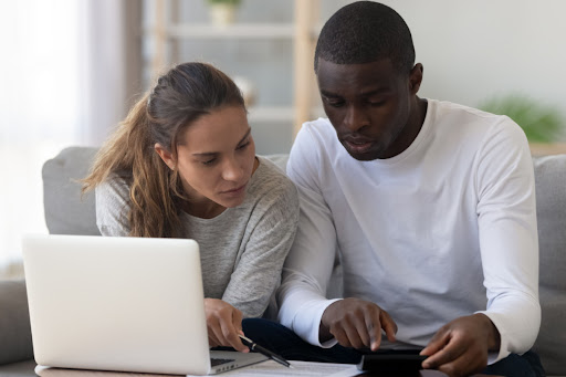 A man and woman pointing at a calculator as they sit on a couch in front of a laptop.