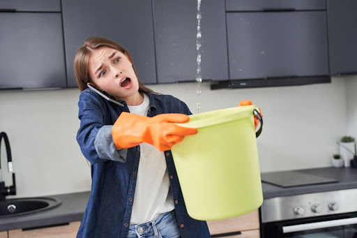 A woman on the phone as she tries to collect leaking water with a bucket.